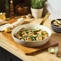 The taupe 12.25-Inch Forged Induction Frying Pan is shown in a lifestyle setting, resting on a wooden cutting board. The pan is filled with a Mediterranean-style sauté of shrimp, sliced zucchini, peas, and charred lemon rounds. In the background, a light-colored kitchen counter holds a potted herb plant, garlic cloves, and several glass bottles.