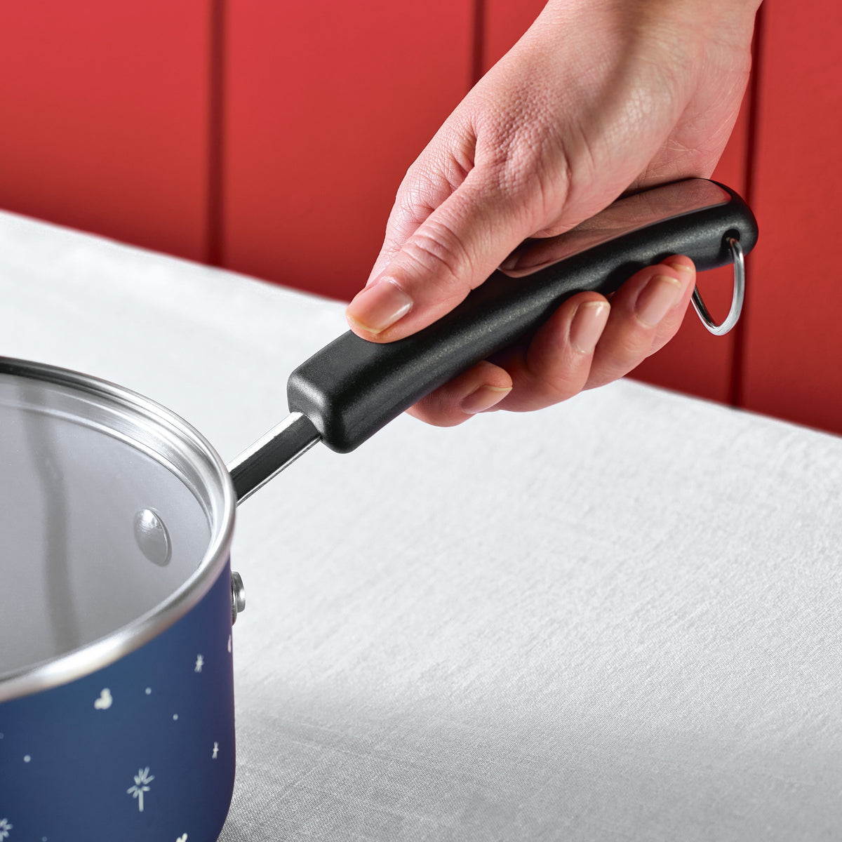 Hand holding a saucepan with a black handle on a white tablecloth.