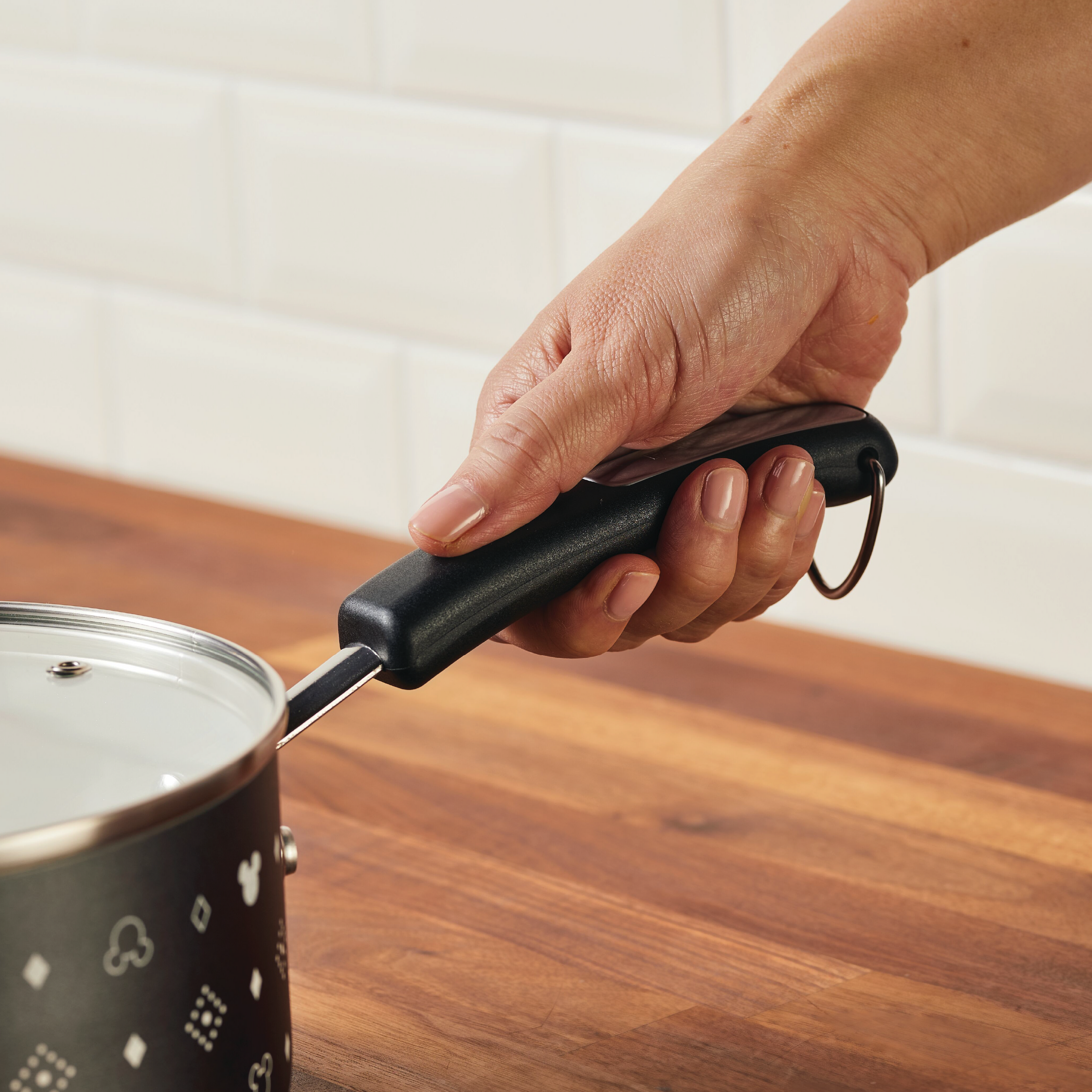 A hand holds a 2-Quart Monochrome Ceramic Nonstick Saucepan with Lid, featuring a Mickey Mouse theme, seamlessly resting on a wooden countertop.