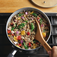 In the Monochrome Collection, a 2-Quart Monochrome Ceramic Nonstick Saucepan with Lid from Farberware cookware sits on the stovetop, filled with quinoa and mixed vegetables, gently stirred with a wooden spoon.