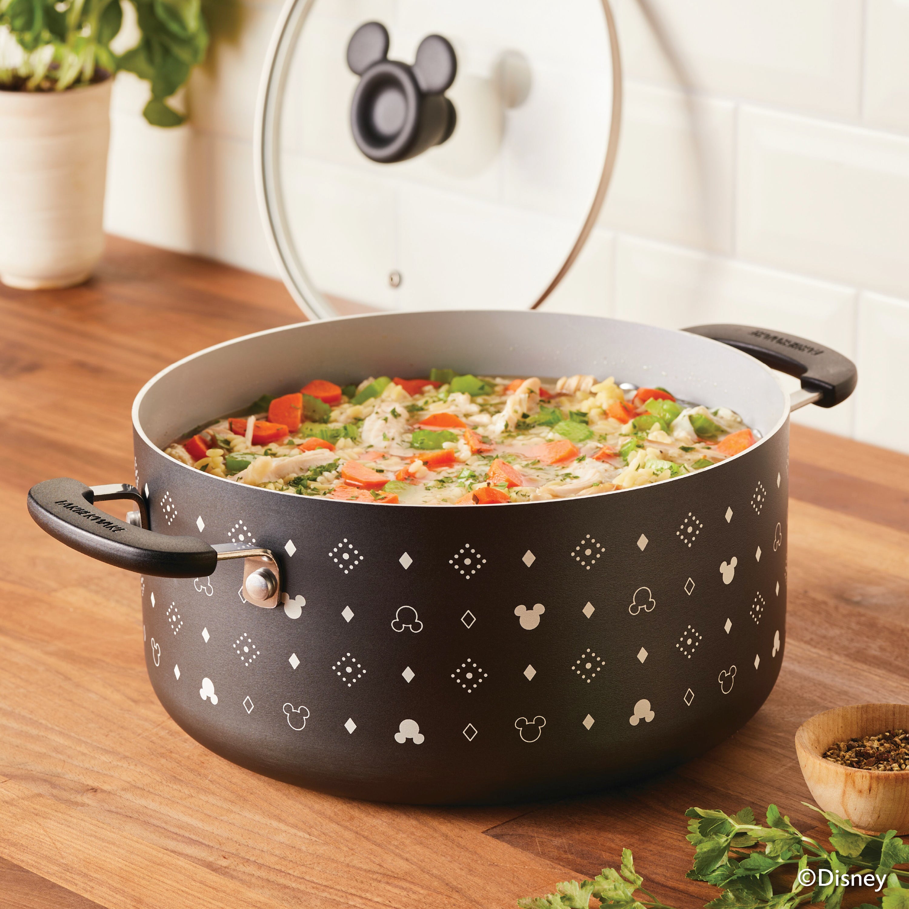 A 5.5-quart monochrome ceramic nonstick stockpot with lid, adorned with Mickey Mouse designs, simmers soup on a wooden countertop.