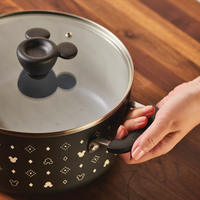 A person holds a 5.5-Quart Monochrome Ceramic Nonstick Stockpot with Lid, adorned with a whimsical Mickey Mouse-themed design, set upon a wooden surface.