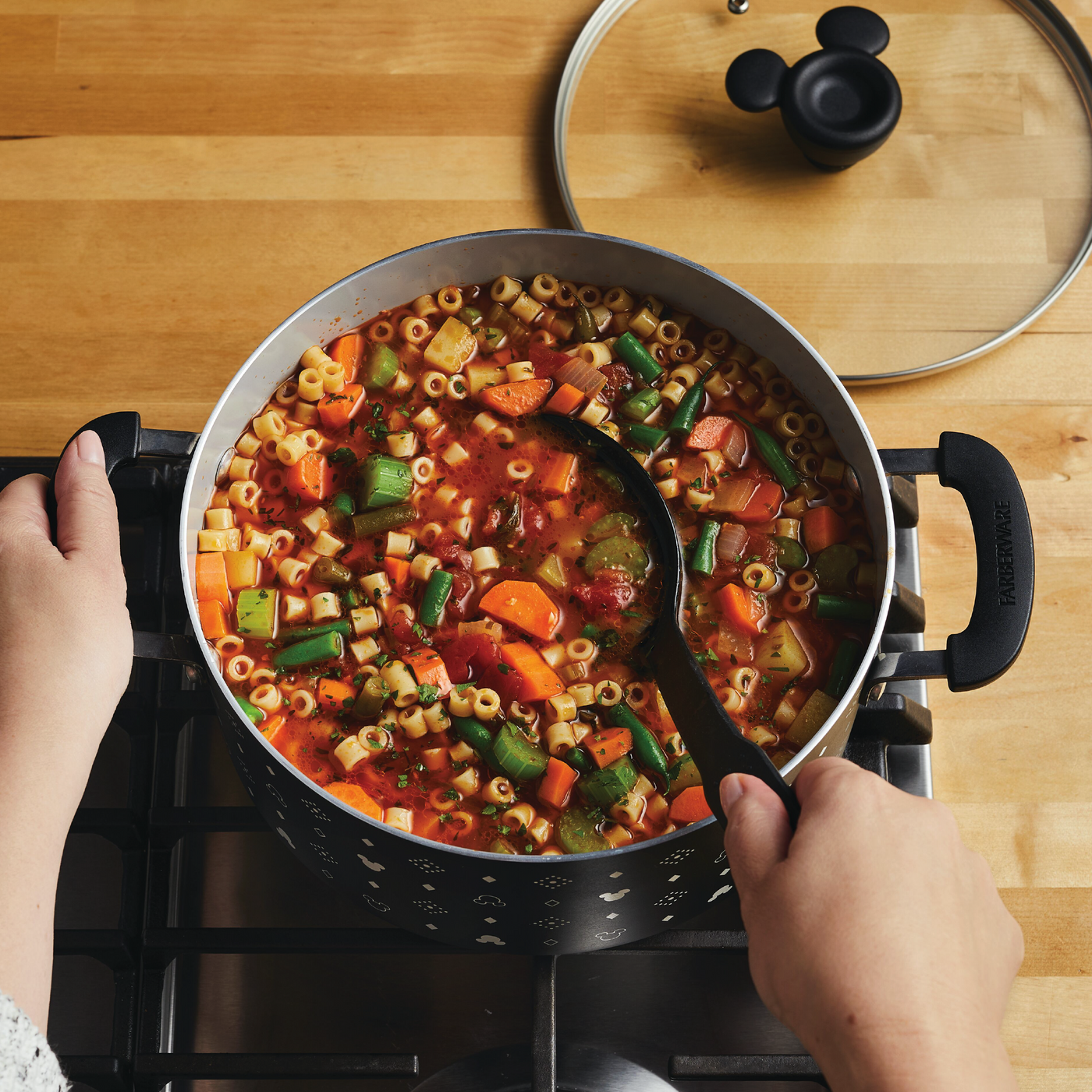 A person stirring a 5.5-Quart Monochrome Ceramic Nonstick Stockpot with Lid filled with vegetable pasta soup on the stovetop, adorned in a whimsical Mickey Mouse apron that adds a touch of Disney magic.