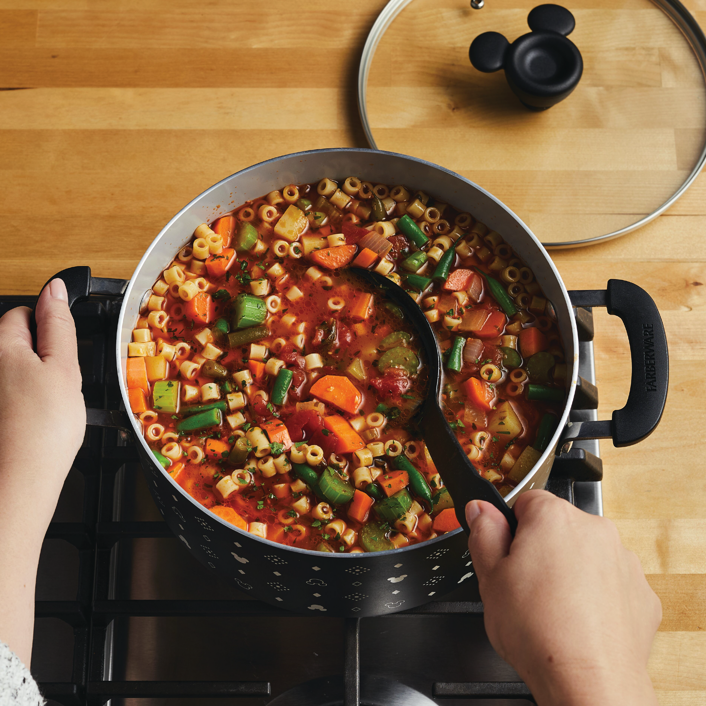 A person stirring a 5.5-Quart Monochrome Ceramic Nonstick Stockpot with Lid filled with vegetable pasta soup on the stovetop, adorned in a whimsical Mickey Mouse apron that adds a touch of Disney magic.