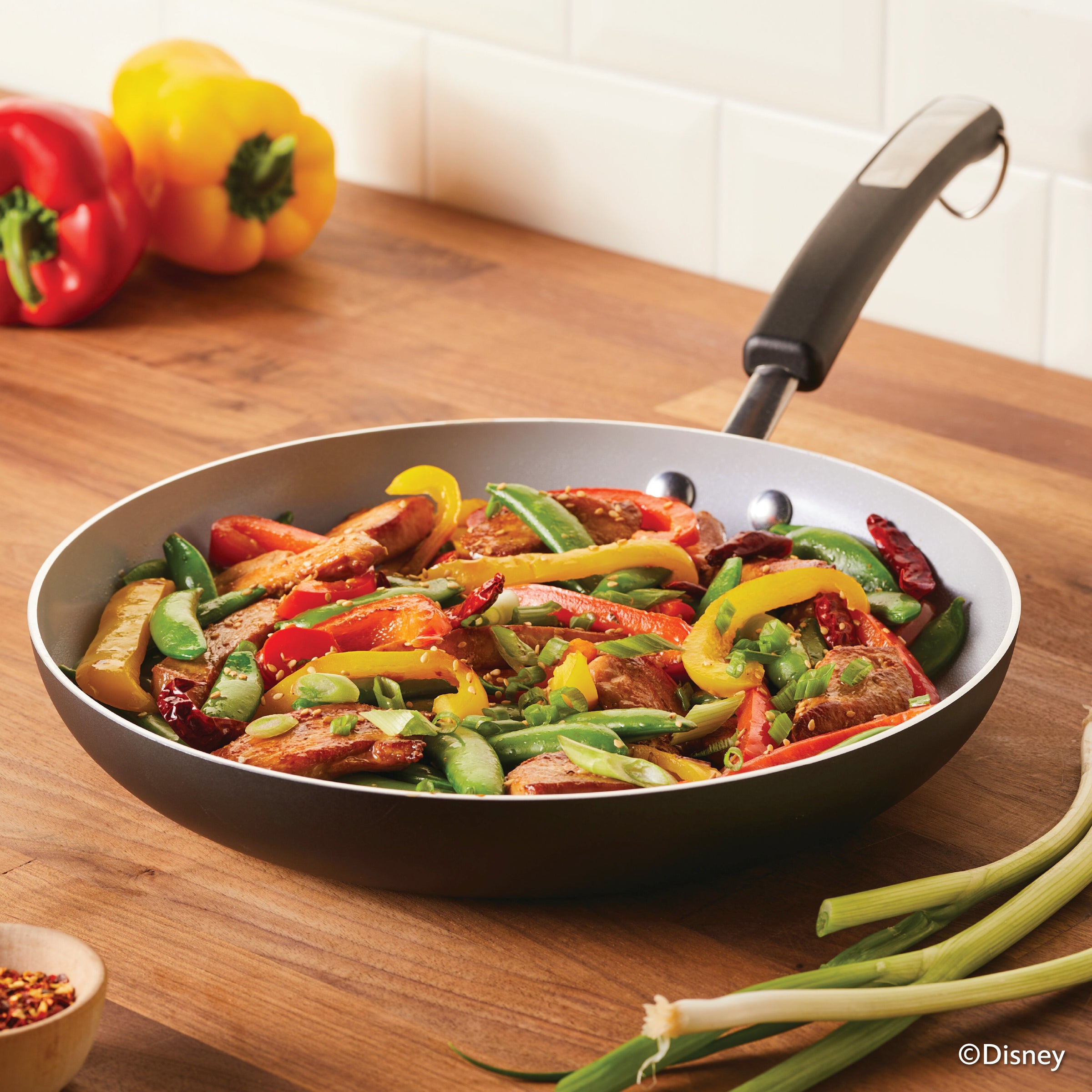 A stir-fry with vegetables and chicken sizzles in a Monochrome Ceramic Nonstick Frying Pan, resting on a wooden surface.