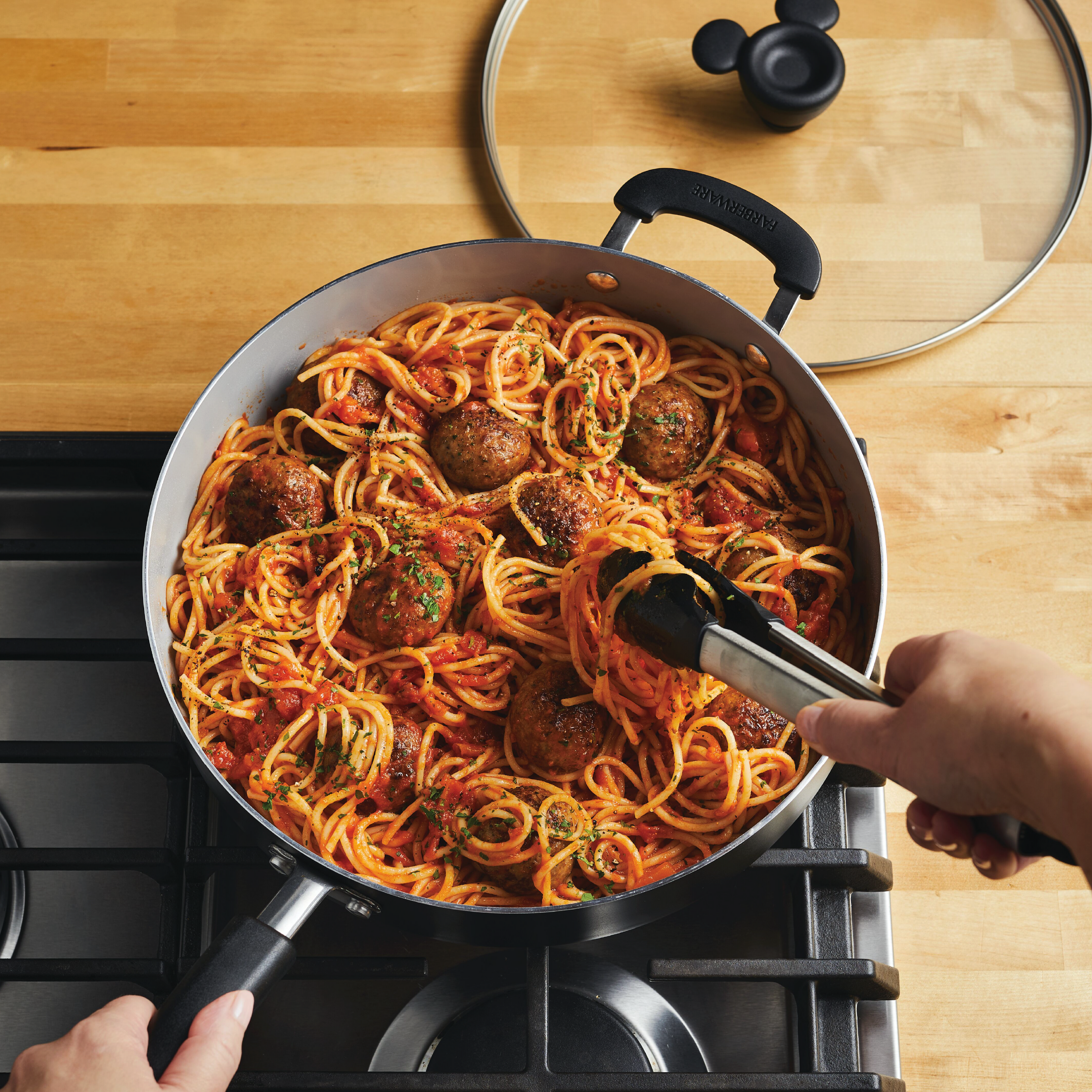A person is stirring spaghetti and meatballs in the 4.5-Quart Monochrome Ceramic Nonstick Sauté Pan with lid and Helper Handle on a stovetop, showcasing the classic elegance of the Monochrome Collection.