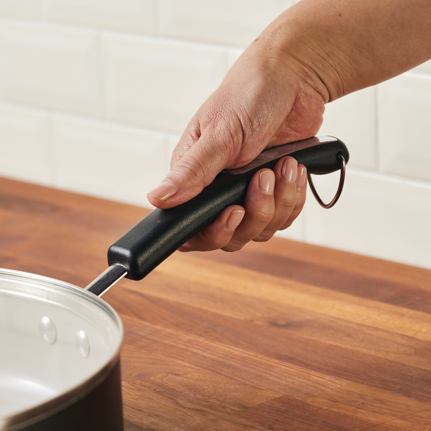 A hand grasps the handle of a 4.5-Quart Monochrome Ceramic Nonstick Sauté Pan with lid and helper handle, resting on a wooden kitchen countertop.