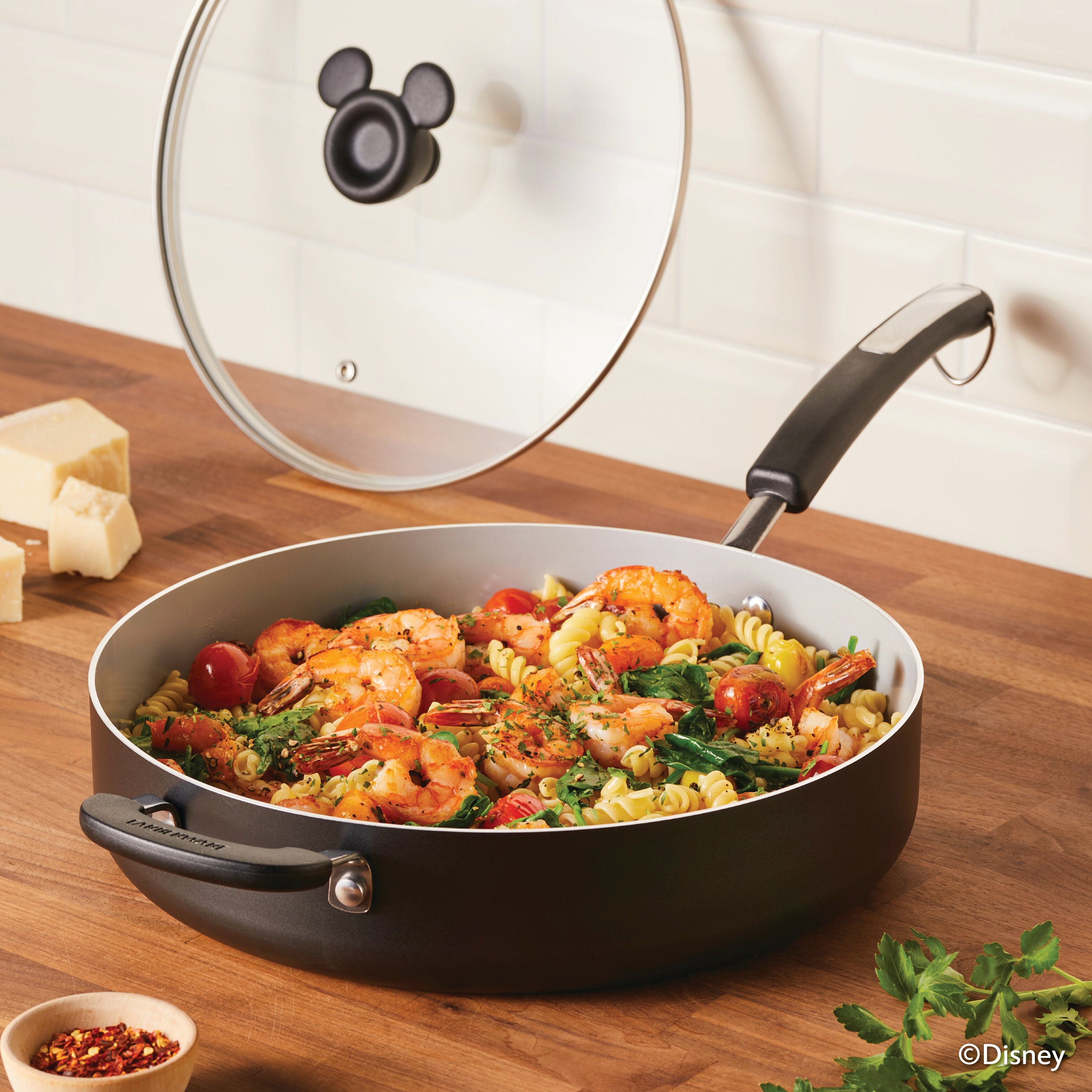 A sleek 4.5-quart Monochrome Ceramic Nonstick Sauté Pan with lid and helper handle sits on the stove, simmering shrimp pasta beneath its clear lid adorned with a charming Mickey Mouse handle.