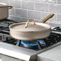 The Radiant Nonstick 2.75-Quart Saute Pan with Lid, featuring a gold accent handle, sits on a lit gas stove burner, ensuring even heat distribution for perfect cooking results.