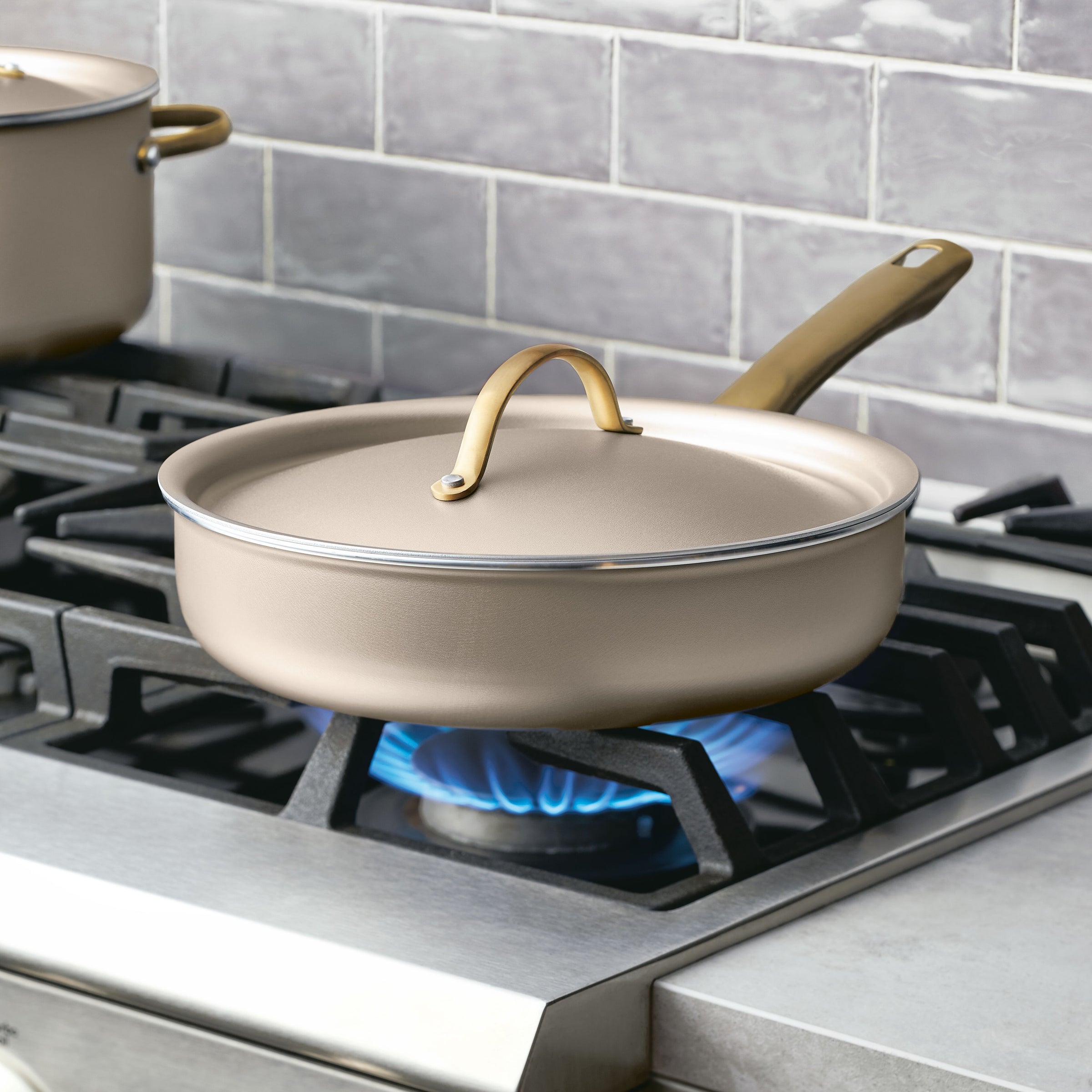 The Radiant Nonstick 2.75-Quart Saute Pan with Lid, featuring a gold accent handle, sits on a lit gas stove burner, ensuring even heat distribution for perfect cooking results.