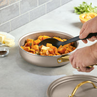 Using a black spoon, a person stirs perfectly cooked squash in the Radiant Nonstick 2.75-Quart Saute Pan With Lid, appreciating its even heat distribution and stylish gold accent handle.