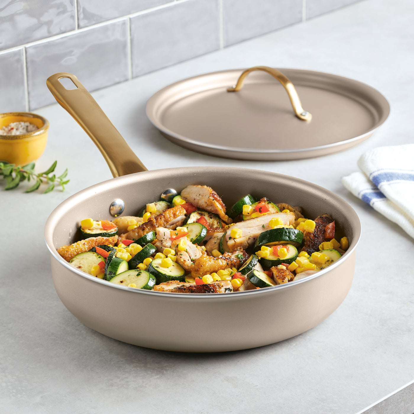 A Radiant Nonstick 2.75-Quart Sauté Pan with Lid, featuring a gold accent handle, sits on the countertop, evenly distributing heat to perfectly cook chicken, zucchini, corn, and tomatoes.