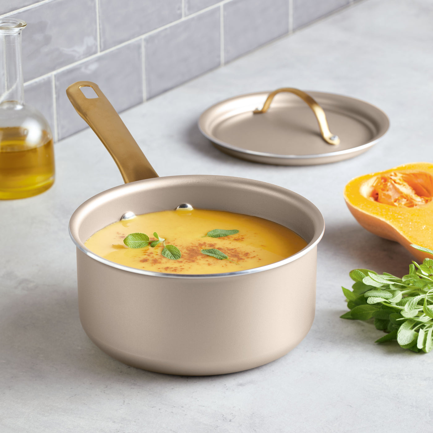 A Radiant Nonstick 2-Quart Saucepan with Lid filled with soup sits beside herbs and a cut pumpkin on the kitchen counter, promising even heat distribution for the perfect meal.