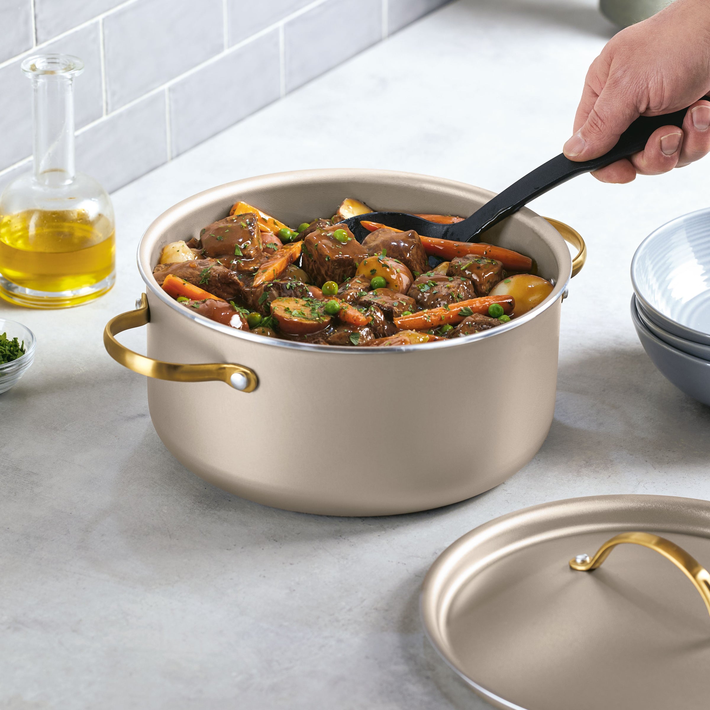 A hand stirs beef stew in the Radiant Nonstick 5.5-Quart Dutch Oven With Lid on a kitchen counter, ensuring even heat distribution for perfect cooking.