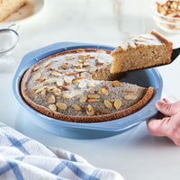 A person serves a slice of almond cake from a blue pan.