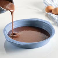 Pouring chocolate batter into a blue round cake pan on a white countertop.