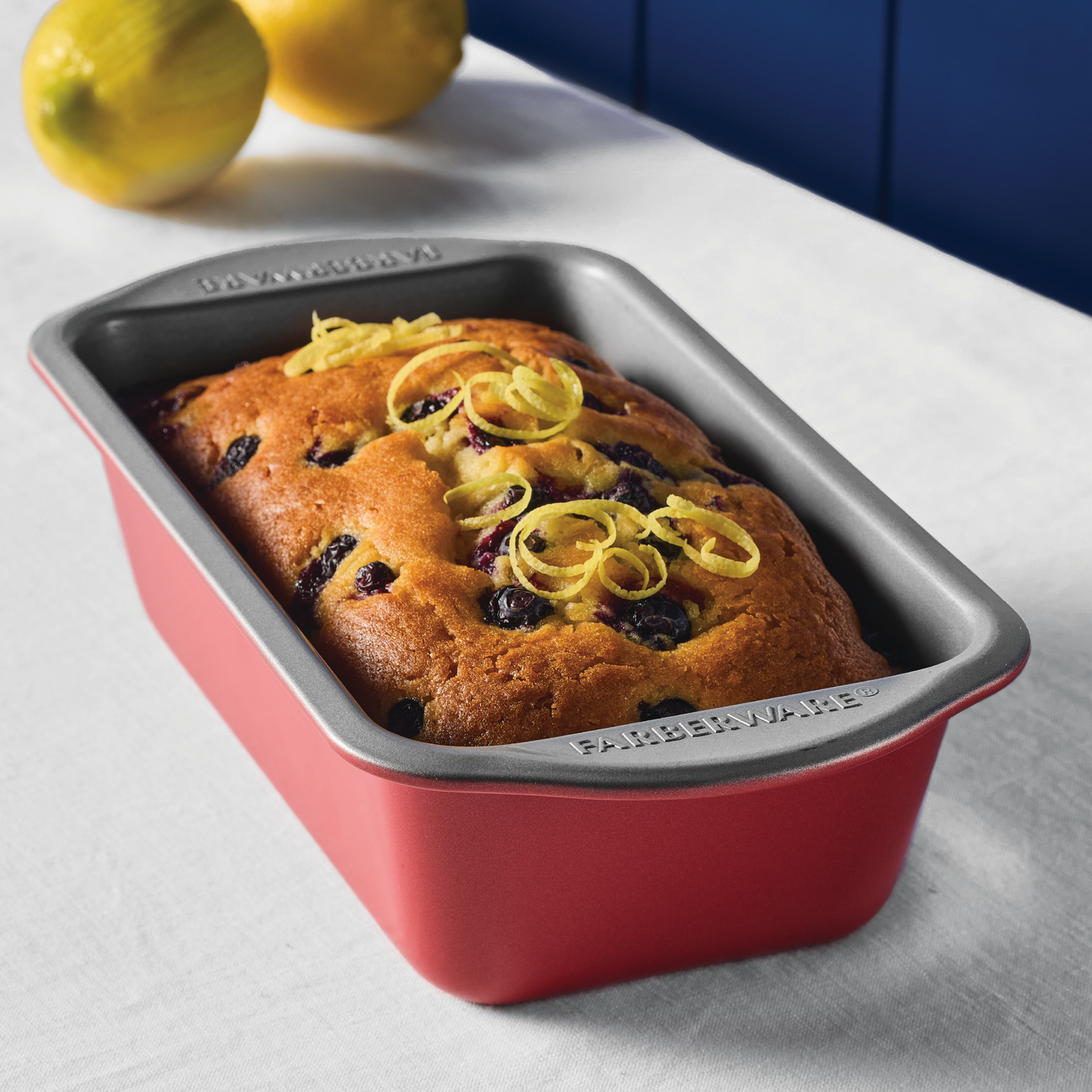 A delightful lemon blueberry loaf rests in a vibrant *9"x5" Nonstick Loaf Pan* on a crisp white tablecloth, showcasing the charm of quality bakeware.