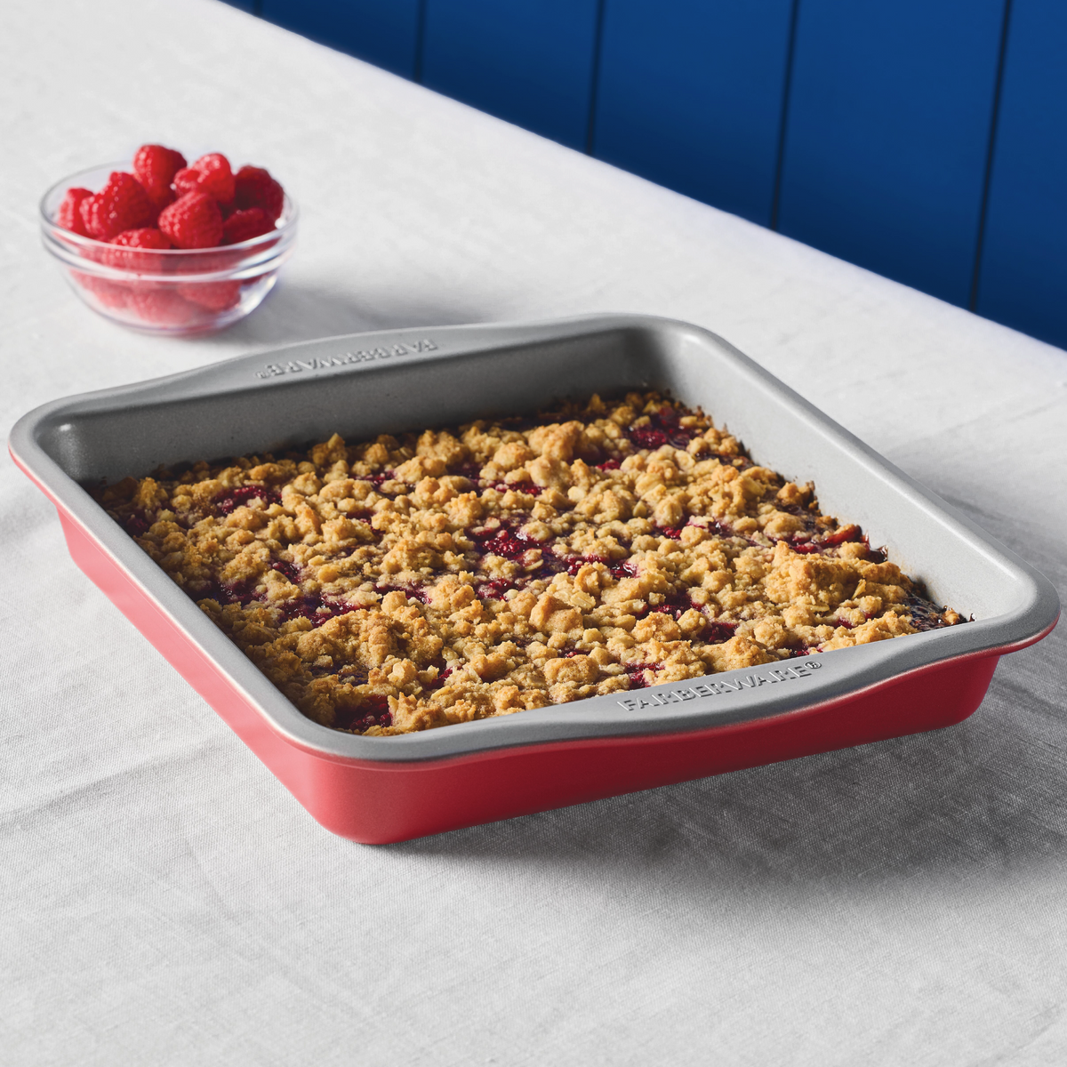 A baked crumble in a red pan next to a bowl of raspberries on a table.