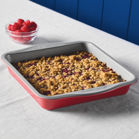 A baked crumble in a red pan next to a bowl of raspberries on a table.