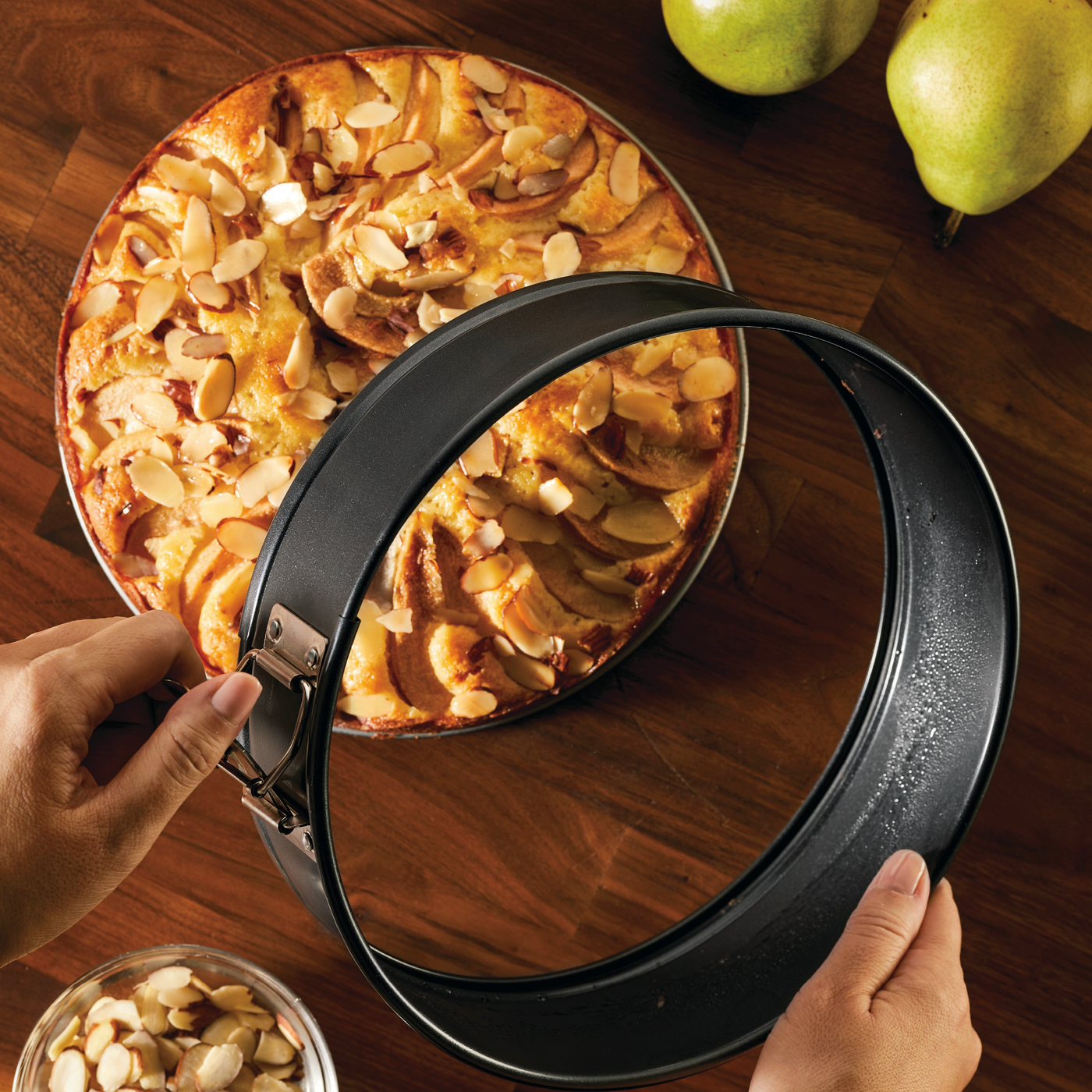 A person expertly removes a 9-Inch Nonstick Round Springform Pan from an almond-topped cake on a wooden surface, demonstrating their proficiency in using bakeware. Bake with Mickey and create delightful treats like this one.