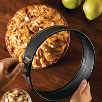 A person expertly removes a 9-Inch Nonstick Round Springform Pan from an almond-topped cake on a wooden surface, demonstrating their proficiency in using bakeware. Bake with Mickey and create delightful treats like this one.
