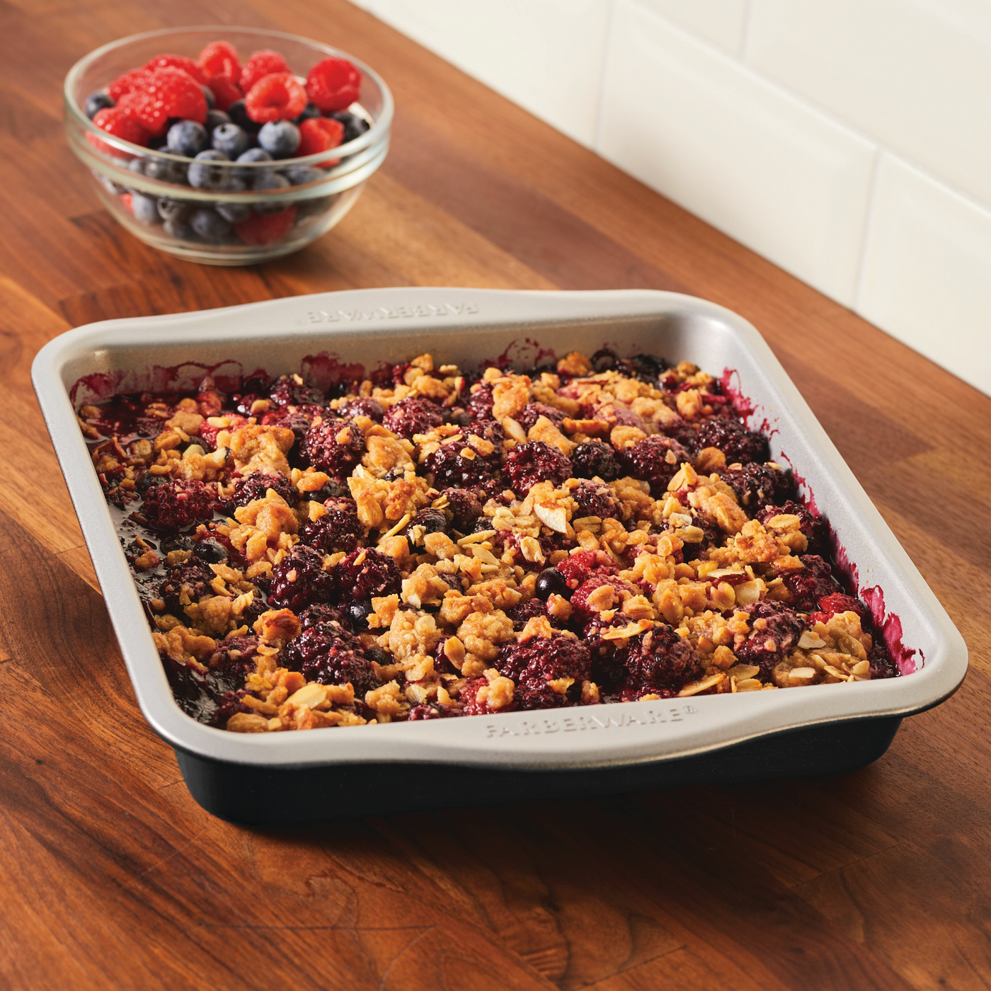 Baking in a nonstick square cake pan on a wooden countertop, bowl of berries behind. Perfect for those who love to bake with Mickey!