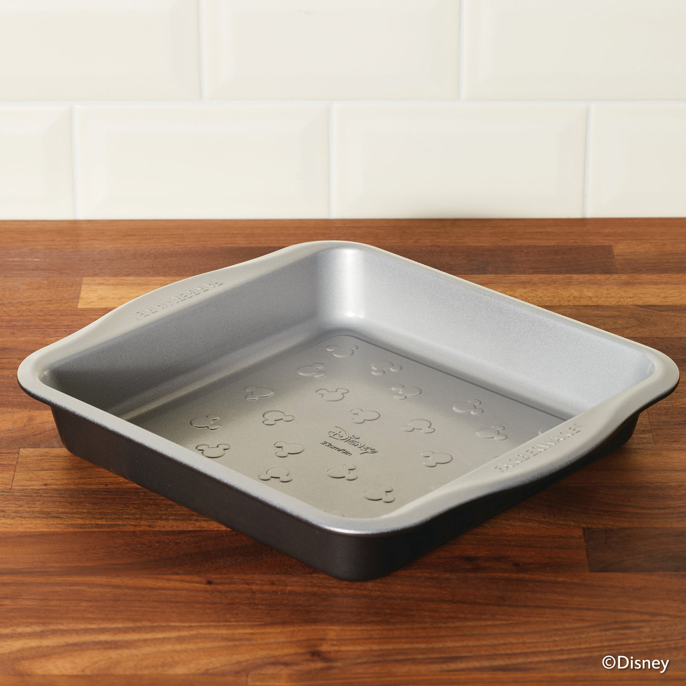 A 9-inch nonstick square cake pan featuring Mickey Mouse patterns rests on a wooden surface, ideal for baking with Mickey in style.