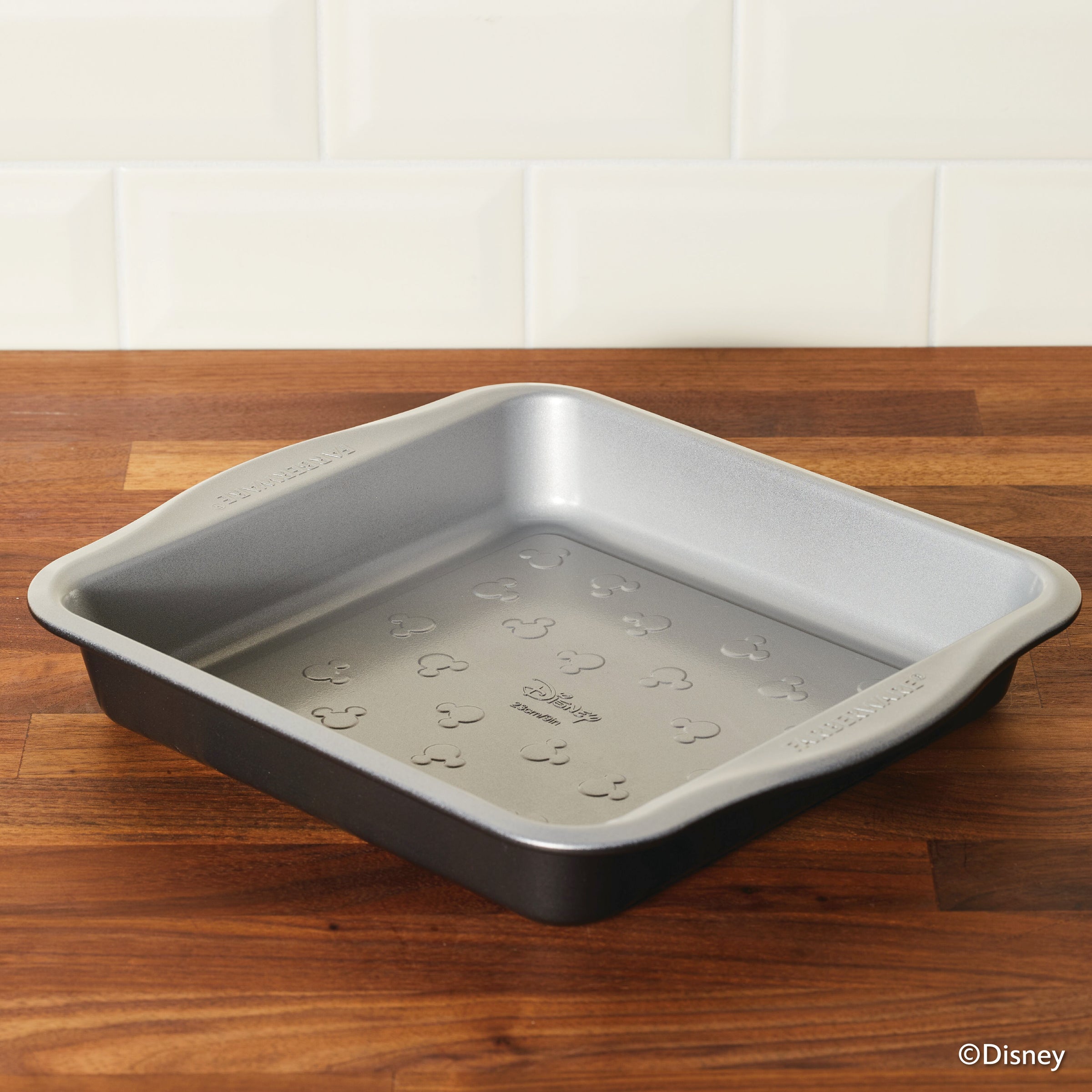 A 9-inch nonstick square cake pan featuring Mickey Mouse patterns rests on a wooden surface, ideal for baking with Mickey in style.