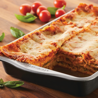 A baked lasagna in a 9" x 13" Nonstick Cake Pan with a slice removed, garnished with tomatoes and basil, makes for the perfect centerpiece to any "Bake with Mickey" themed gathering.