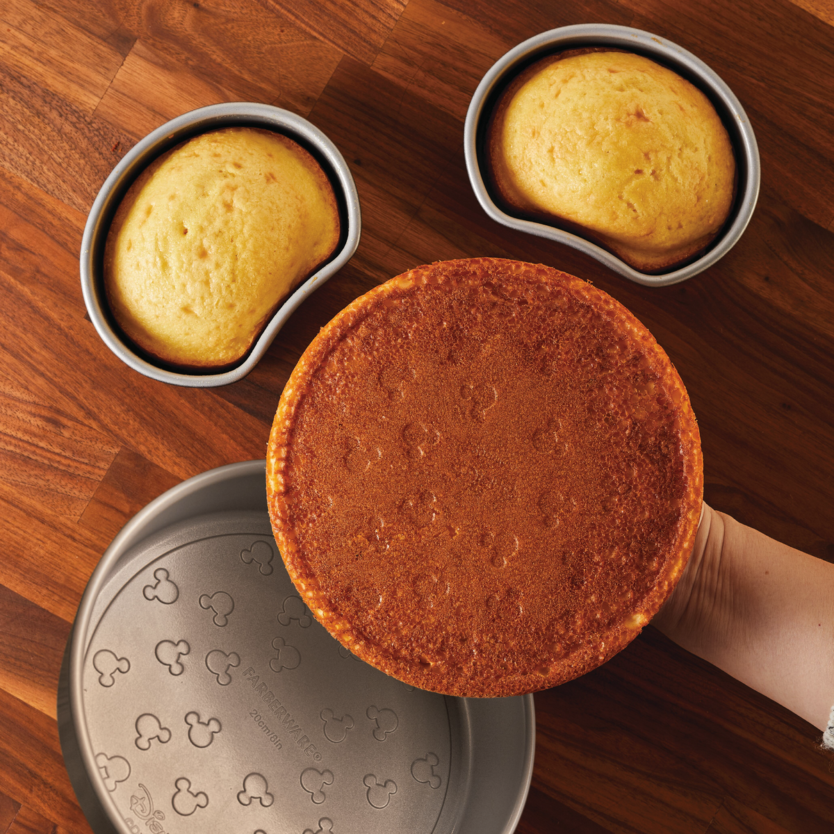 Three baked cakes on wooden surface, one being held by a hand.