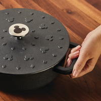A hand holds a pot lid from the 3-Quart Monochrome Pre-Seasoned Cast-Iron Dutch Oven, featuring a charming mouse ear pattern reminiscent of Disney Mickey Mouse cookware, resting on a wooden table.