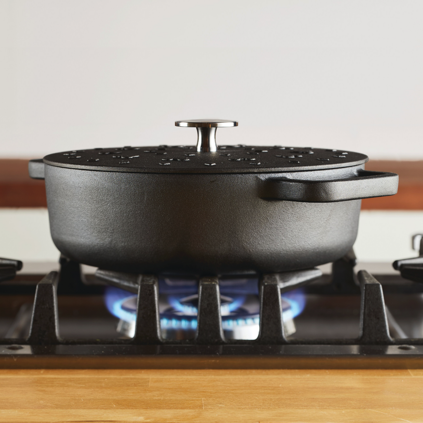 A 3-Quart Monochrome Pre-Seasoned Cast-Iron Dutch Oven from the Disney Mickey Mouse cookware collection sits on a gas stove, its sturdy form silhouetted against the vibrant blue flame.