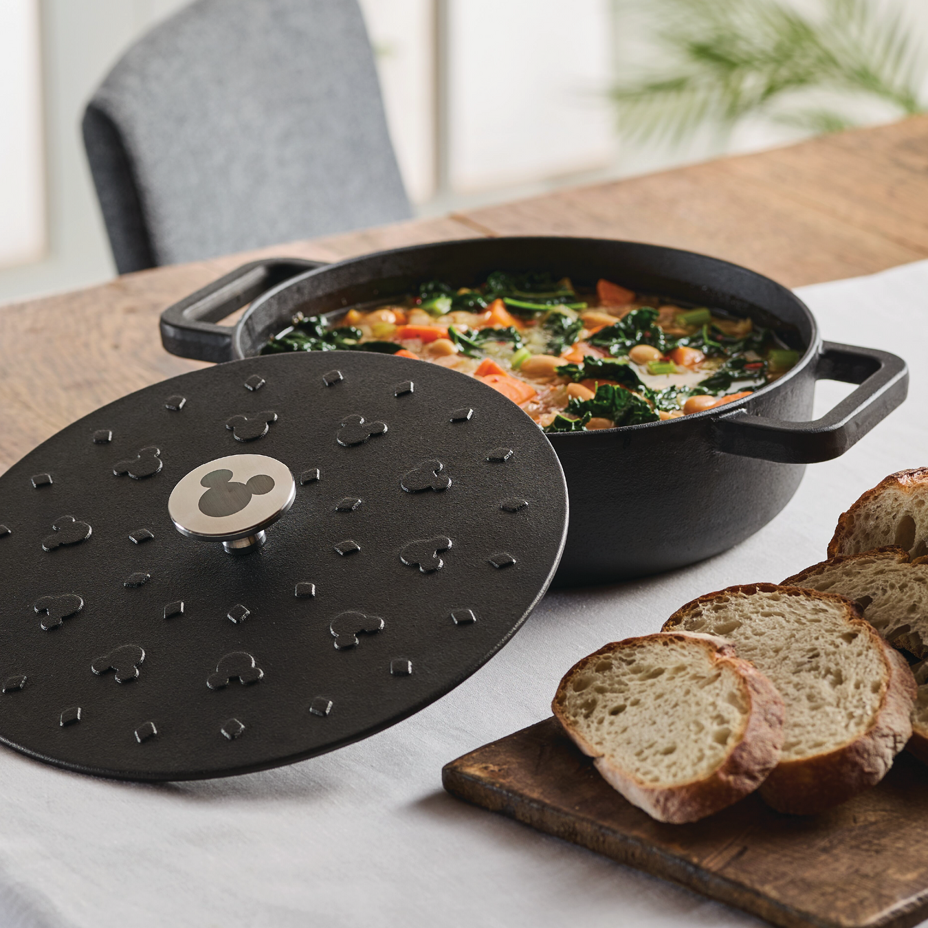 A black 3-Quart Monochrome Pre-Seasoned Cast-Iron Dutch Oven from Farberware is filled with vegetable soup, its lid off, filling the air with inviting aromas across the table. Nearby, freshly sliced bread awaits, completing this cozy culinary scene. product