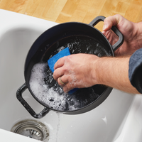 Cleaning a black 3-Quart Monochrome Pre-Seasoned Cast-Iron Dutch Oven from the Disney Mickey Mouse cookware collection with a blue sponge in a white sink.