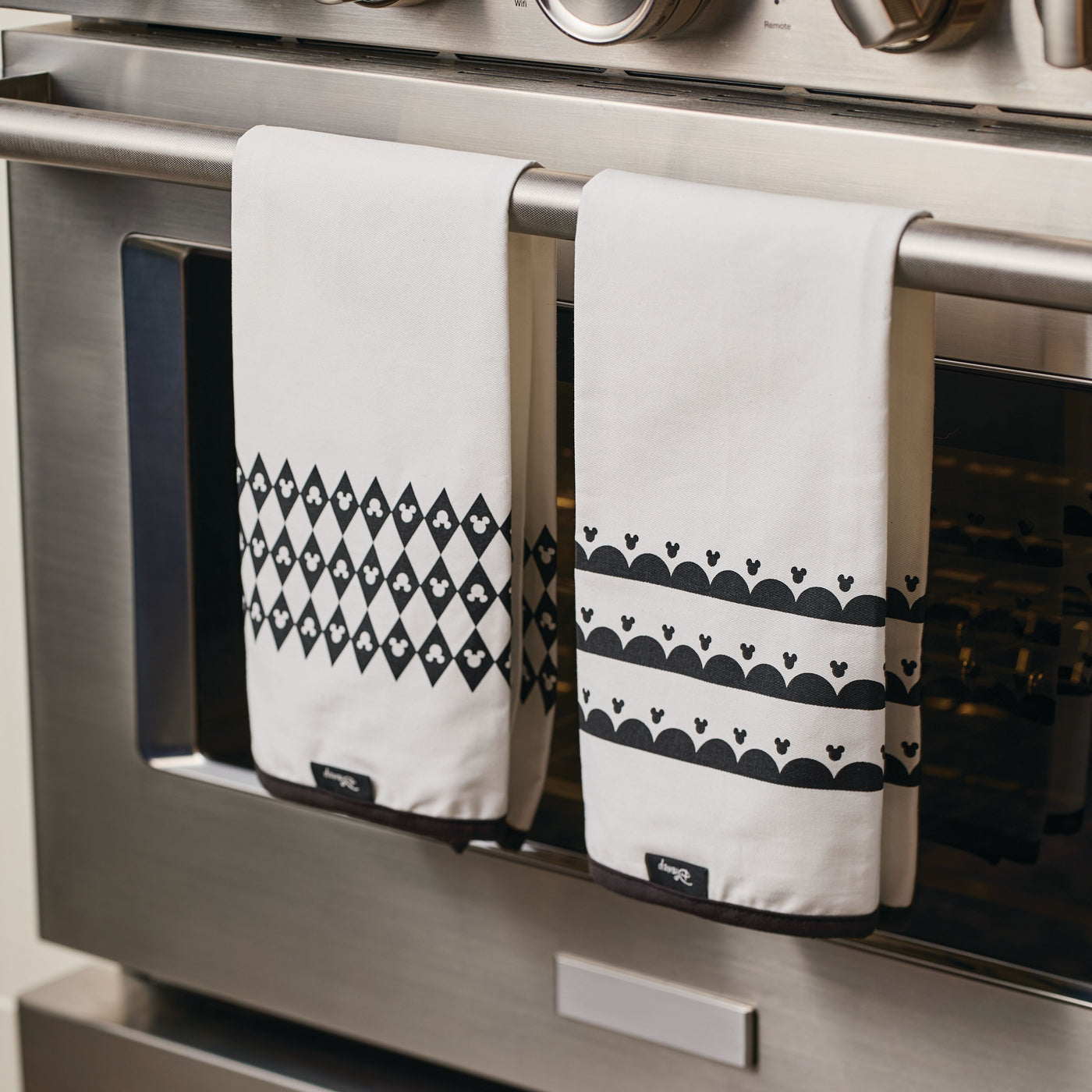 Two stylish Disney "Home Monochrome" kitchen towels, featuring Mickey Mouse-inspired black patterns, are draped elegantly over an oven handle.