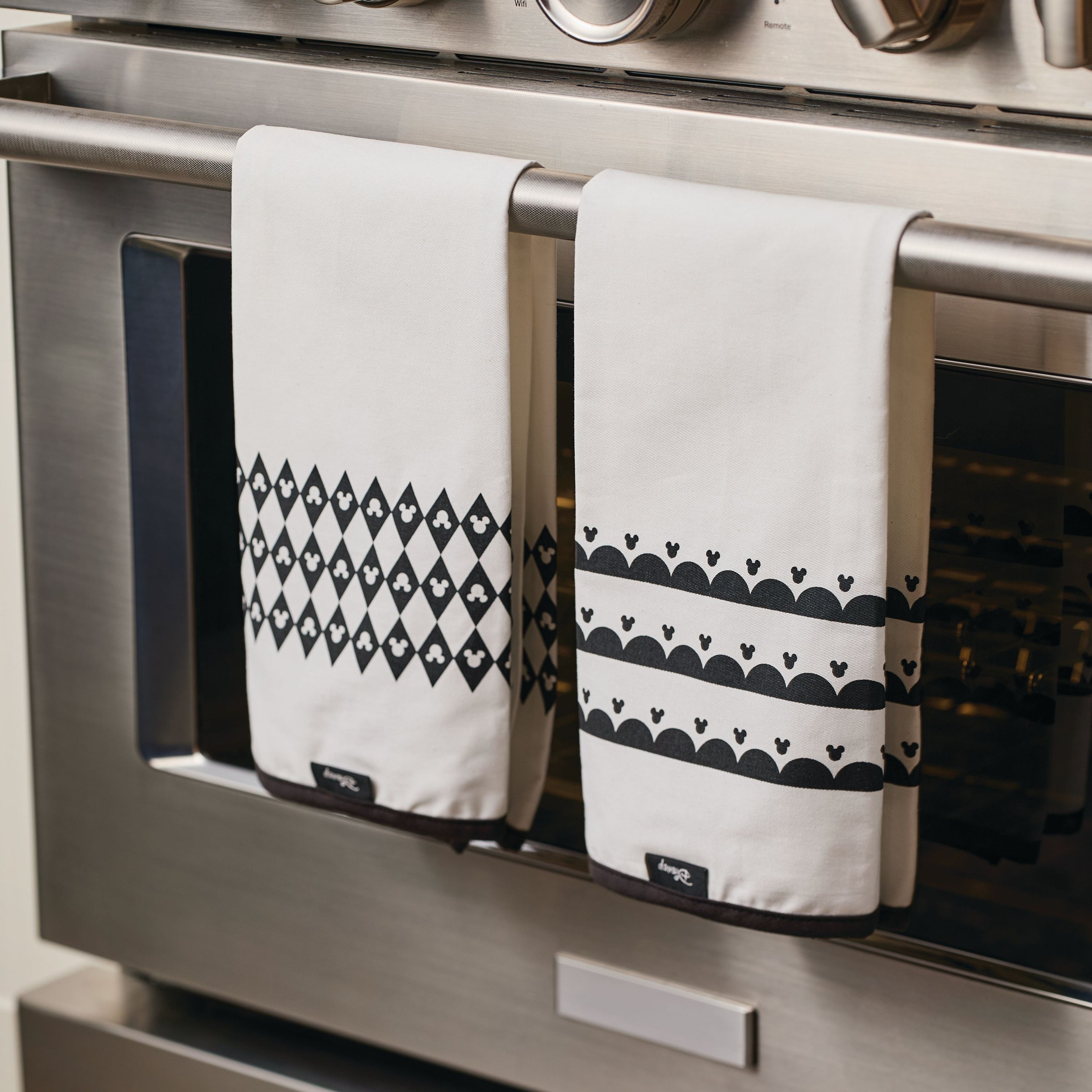 Two stylish Disney "Home Monochrome" kitchen towels, featuring Mickey Mouse-inspired black patterns, are draped elegantly over an oven handle.