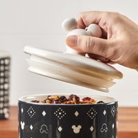 A hand opens a jar from the 3 Piece Set of Ceramic Jar with Airtight Lids, filled with mixed nuts and dried fruits, adding a whimsical touch to your kitchenware collection.