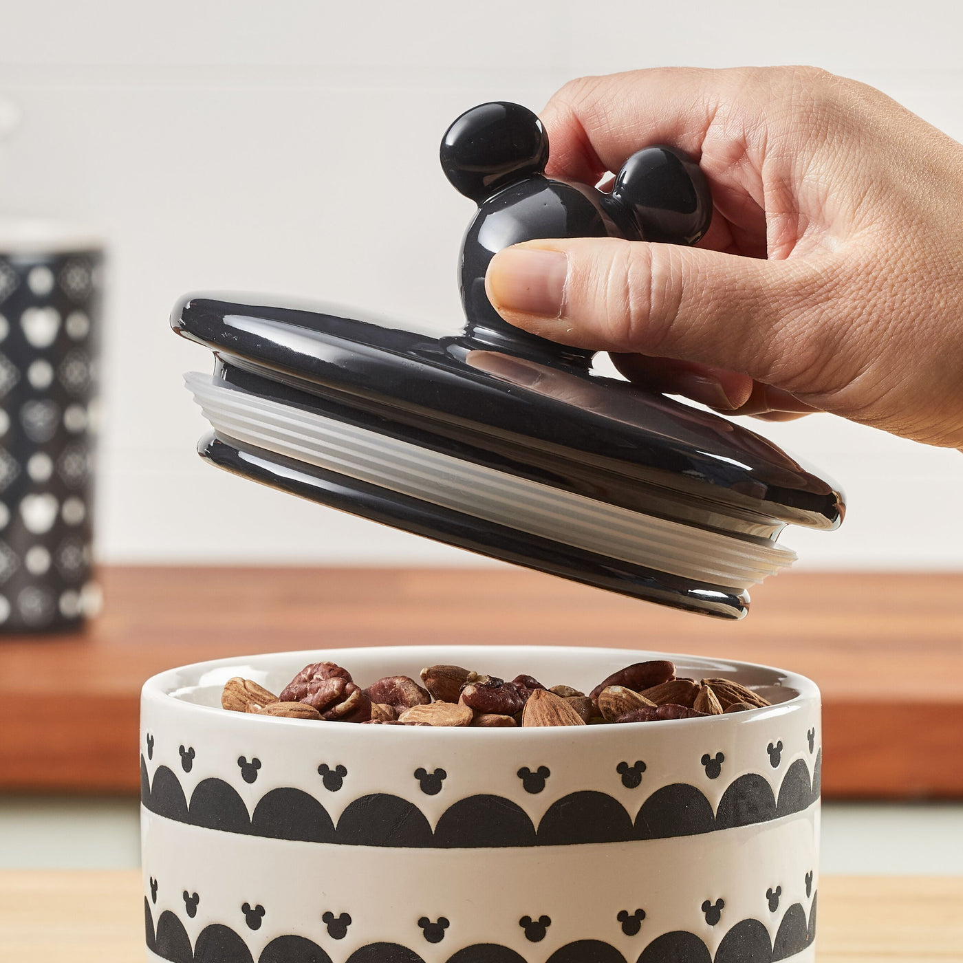 A hand lifting the airtight lid from the Ceramic Jar with Airtight Lid, which is filled with nuts.