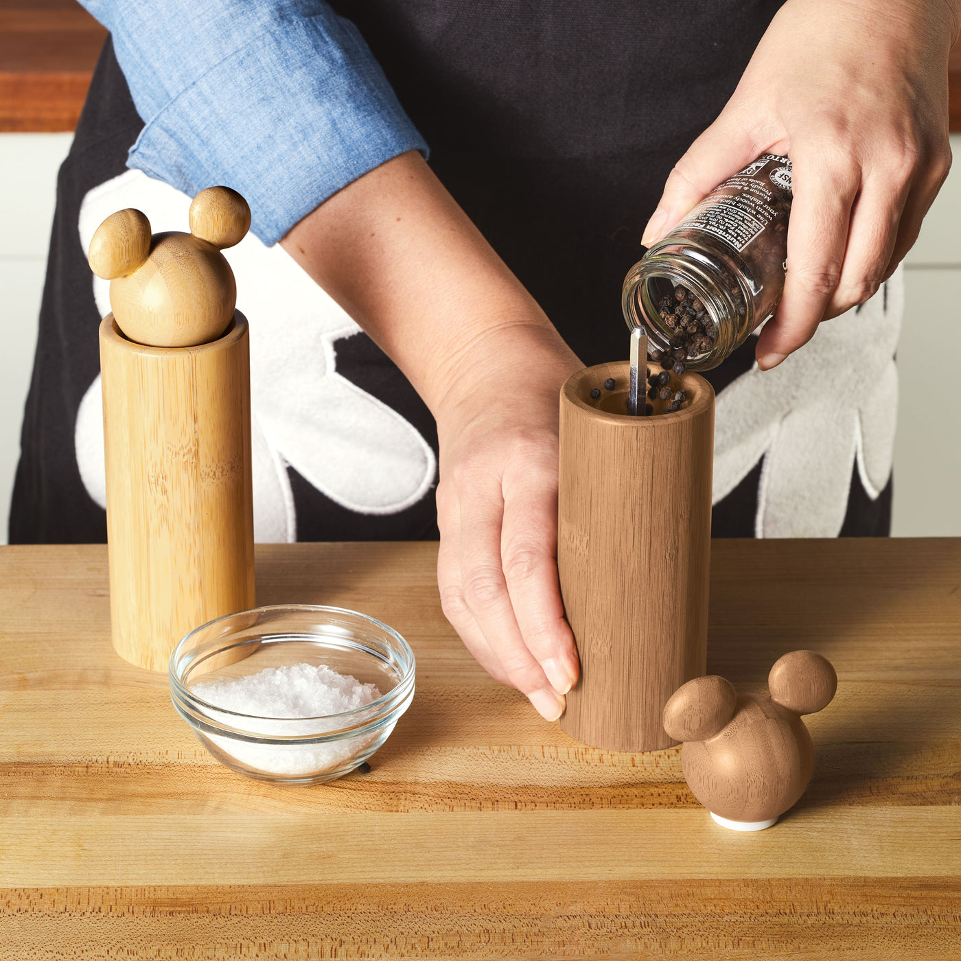 In a kitchen setting, a person fills a charming wooden salt and pepper grinder, reminiscent of the Disney Home collection, adding a touch of whimsy akin to Mickey Mouse's playful spirit.