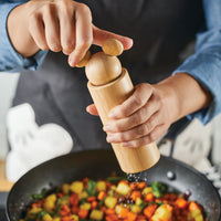 A person is using a wooden salt and pepper grinder with a Mickey Mouse theme over a pan of mixed vegetables.
