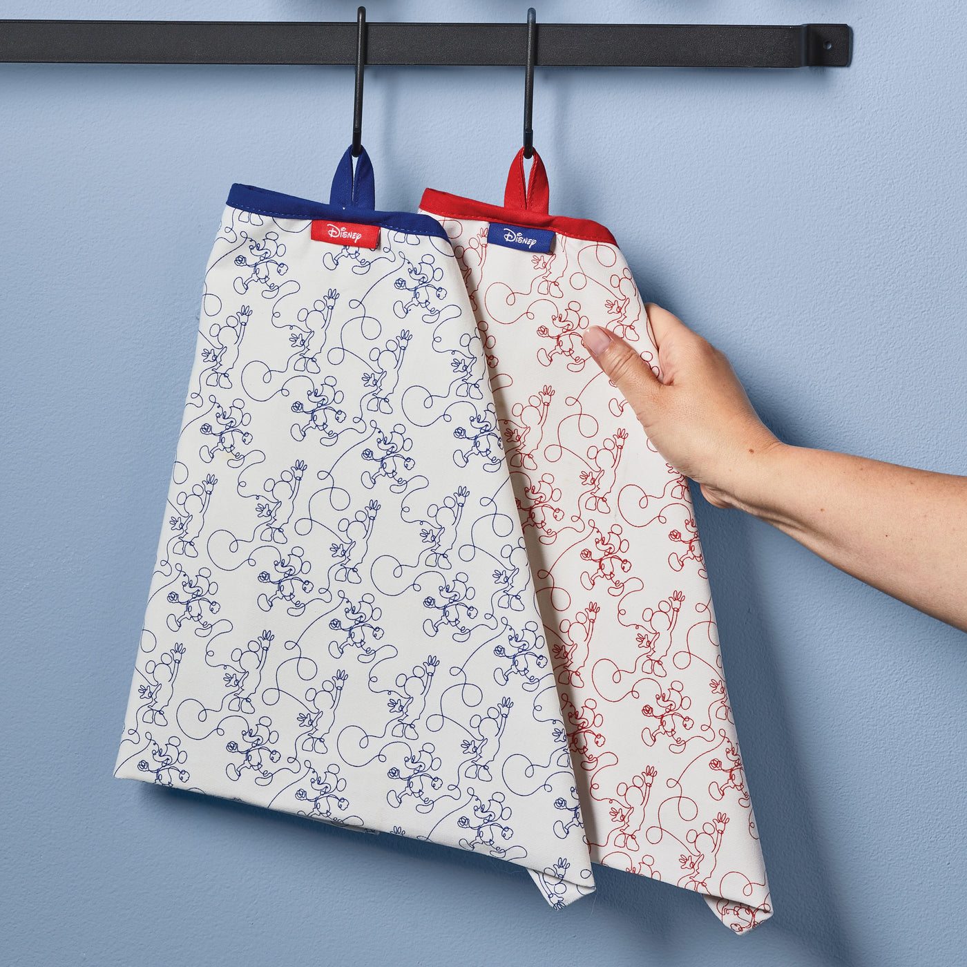 A hand holding two patterned Disney kitchen towels from the Bon Voyage collection on a wall hook.