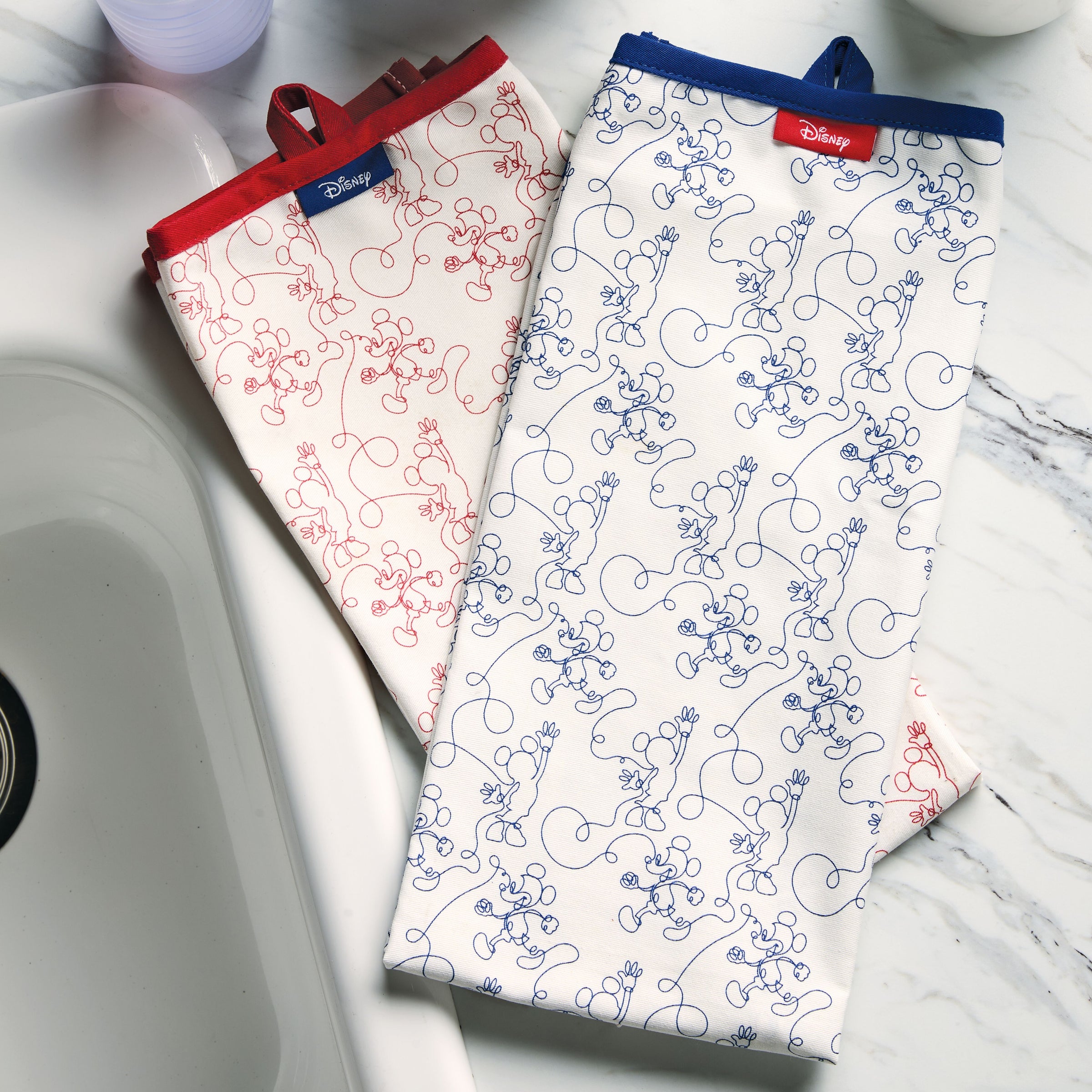 Two Bon Voyage Kitchen Towels featuring Mickey Mouse from Disney rest on a marble countertop next to a sink.