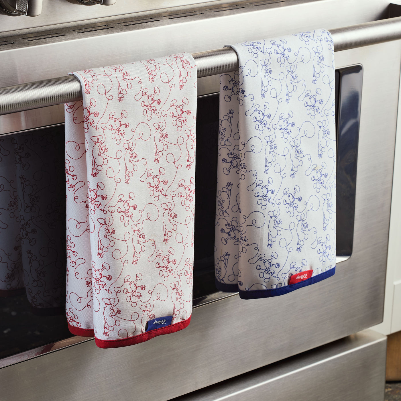 Two Bon Voyage Kitchen Towels, showcasing intricate red and blue designs, hang on an oven handle.