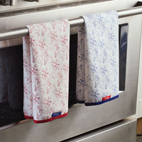 Two Bon Voyage Kitchen Towels, showcasing intricate red and blue designs, hang on an oven handle.