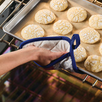 A hand clad in a Bon Voyage Pot Holder retrieves a tray of cookies from the oven, adding a touch of whimsy to your kitchenware ensemble.