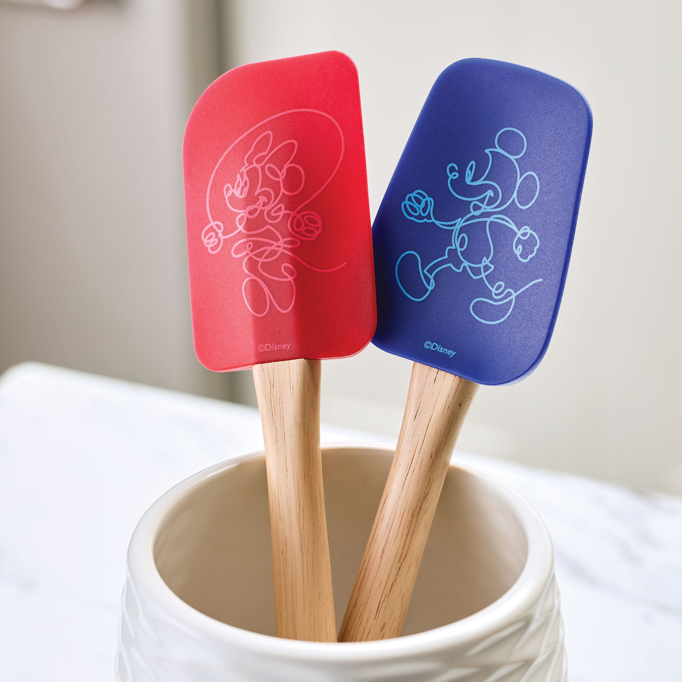 The Bon Voyage 2-Piece Spatula and Spoonula Set, adorned with a charming Disney Mickey Mouse design in red and blue, rests in a white holder on the kitchen counter.