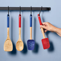 A hand grips the spatula from the Bon Voyage 2-Piece Spatula and Spoonula Set while three wooden kitchen utensils, including the cleverly designed spoonula, hang in the background.