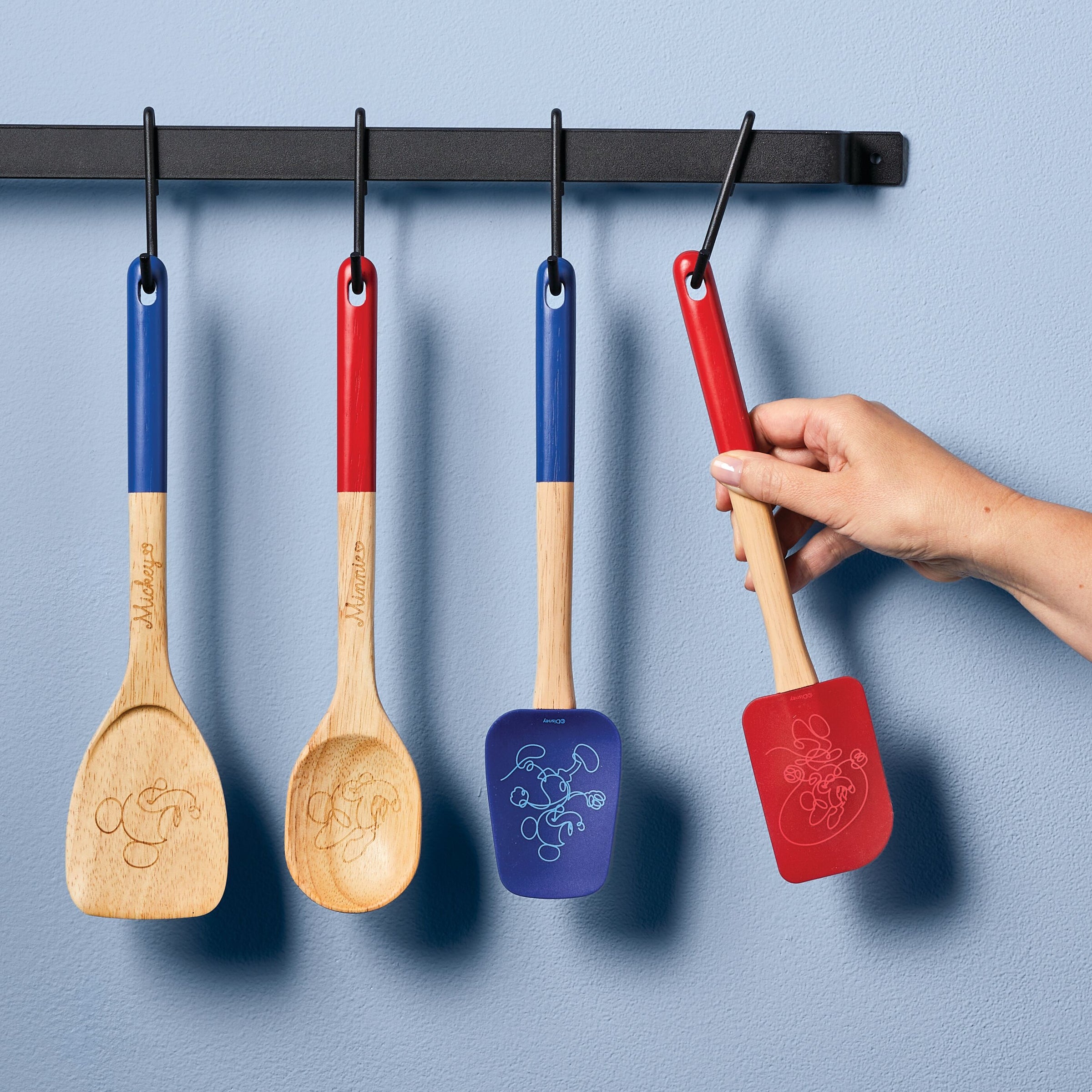 A hand grips the spatula from the Bon Voyage 2-Piece Spatula and Spoonula Set while three wooden kitchen utensils, including the cleverly designed spoonula, hang in the background.
