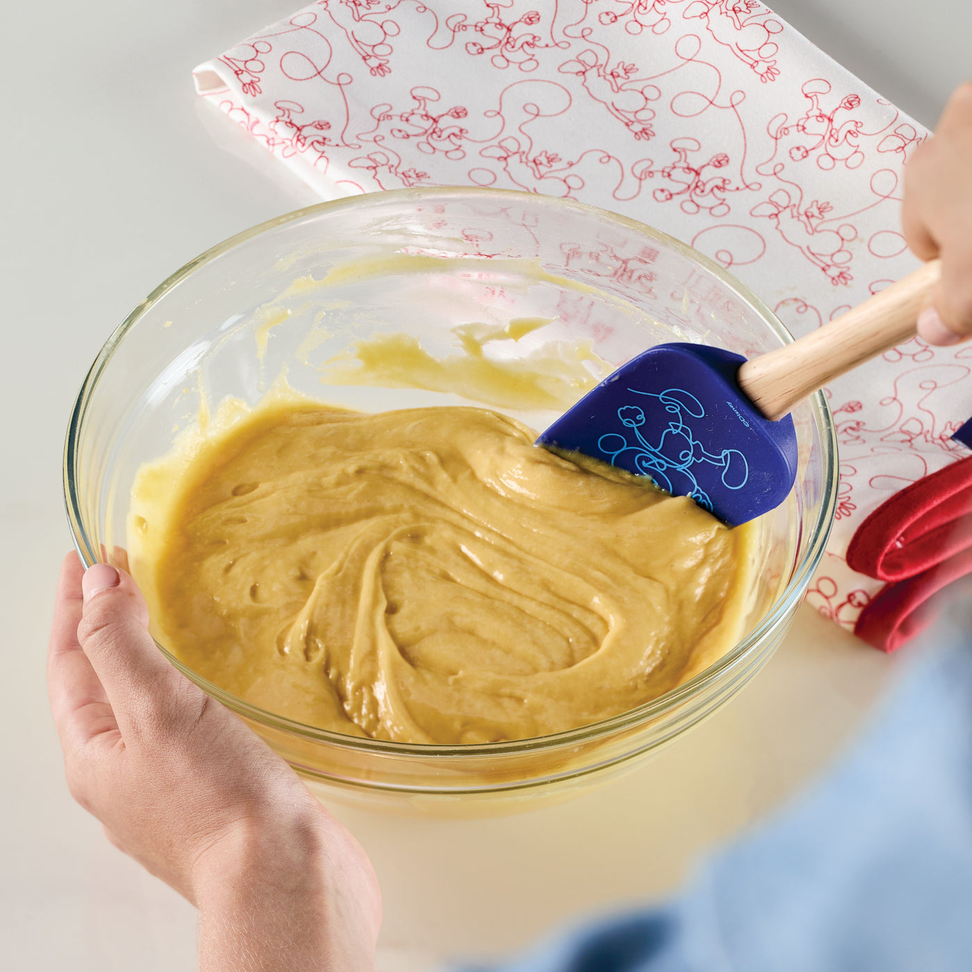 A person is mixing batter in a glass bowl with a blue spatula from the Bon Voyage 2-Piece Spatula and Spoonula Set featuring Disney's Mickey Mouse.