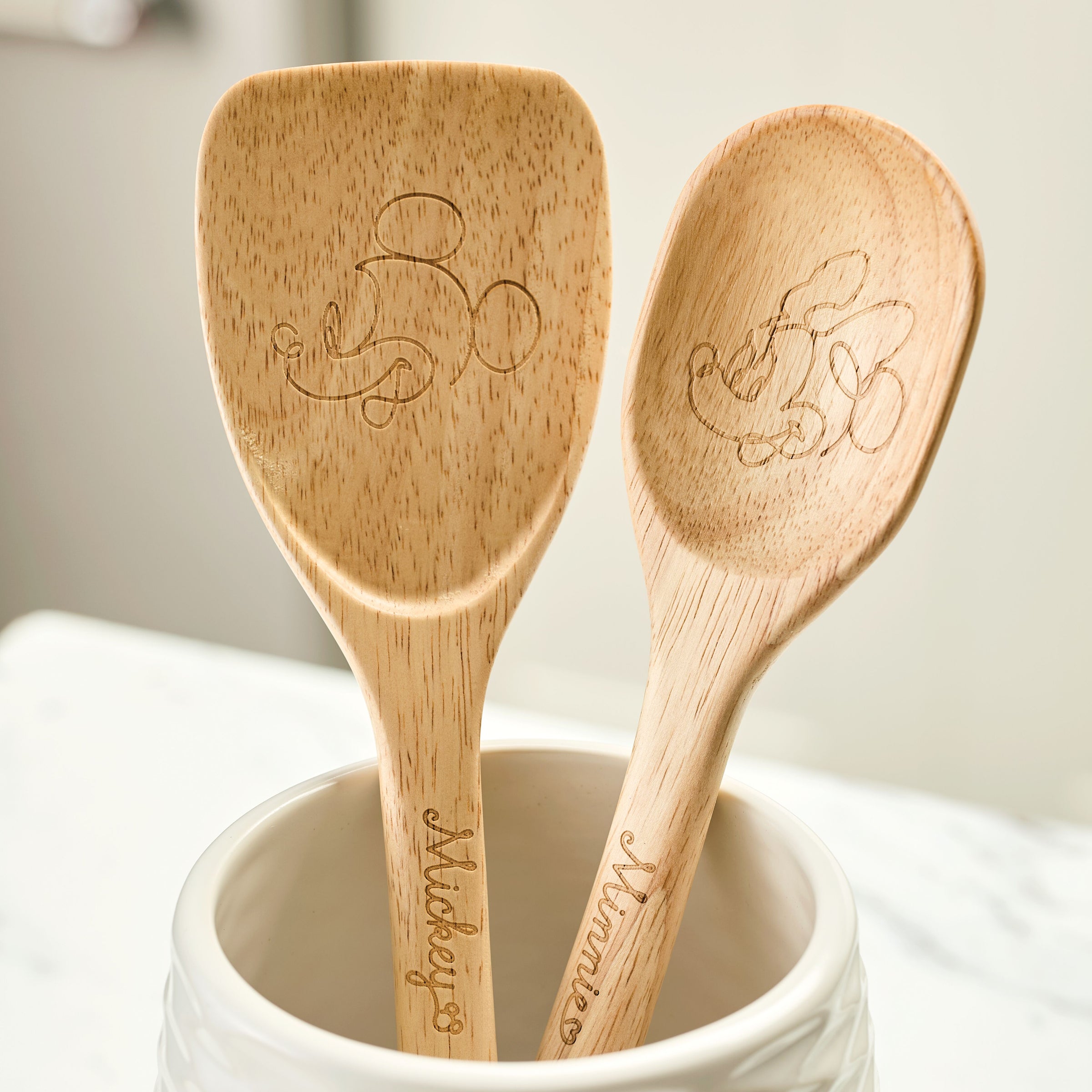 The Bon Voyage 2-Piece Spoon and Turner Set from the Disney Home collection features two wooden cooking spoons engraved with designs of beloved cartoon characters, including Mickey Mouse.